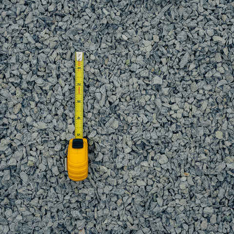 A yellow tape measure is laid vertically on a bed of gray gravel and crushed stone.