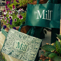 two mill branded tote bags hanging off of plants in a garden