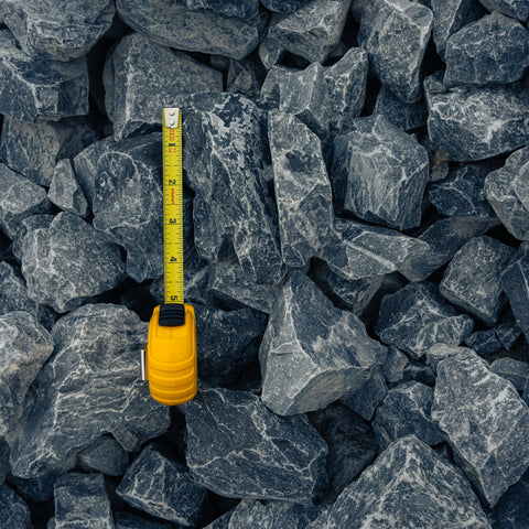 Dark gray angular stone chips or gravel with a yellow tape measure standing upright in the center to show size against the pile.
