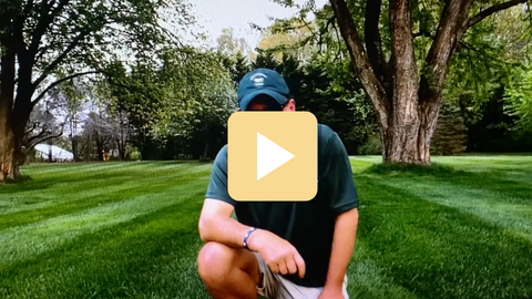 Person in a green shirt and cap sitting on grass with a play button overlay