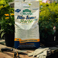 Bag of Bio-tone Organic Starter Plant Food on a wooden surface with gardening tools and plants in the background.