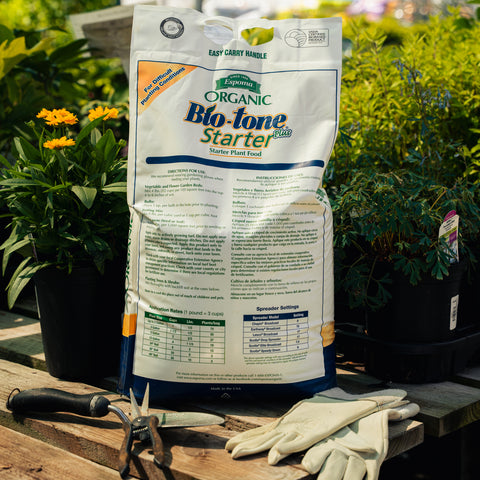 Bag of Bio-tone starter fertilizer on a wooden table with gardening tools and gloves, surrounded by plants.