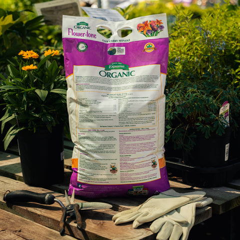 Bag of Espoma Organic flower-tone fertilizer on a wooden surface with gardening tools and plants in the background.