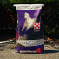 A bag of Purina Ultium Gastric Care horse feed standing outdoors on pavement with dark green foliage in the background. The bag is primarily purple and white with a large photo of a white horse and the Purina logo.