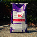 A large bag of Ultium food stands upright outdoors on asphalt with trees and shrubs in the background. The bag is predominantly purple and white with red and black printed panels and text.