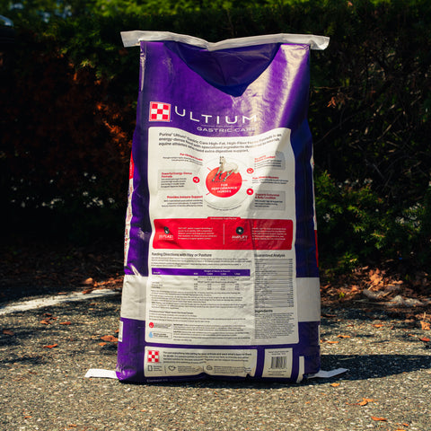 A large bag of Ultium food stands upright outdoors on asphalt with trees and shrubs in the background. The bag is predominantly purple and white with red and black printed panels and text.