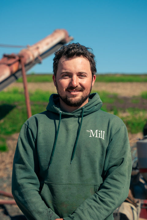 connor mann wearing a mill branded sweatshirt standing near a field on a sunny day