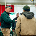 Man in green with a hat talking to another person. 