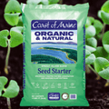 A 16 qt bag of Coast of Maine Seed Starter Organic Seed Starter, featuring a green package with a central illustration of a winding river and forest against a background of newly sprouted plants.