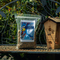 A clear plastic bag of Outdoor Finch Blend birdseed sits on a metal garden table next to a wooden birdhouse, with a decorative metal garden bench and green foliage in the background.