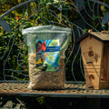 A bag of Mill Chesapeake Birdseed sits on a metal garden bench next to a wooden birdhouse, with green garden foliage in the background.