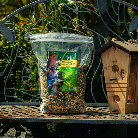 A bag of Mill Backyard Conservation Mix seed mix displayed on a metal garden bench next to a wooden birdhouse, with a garden background.