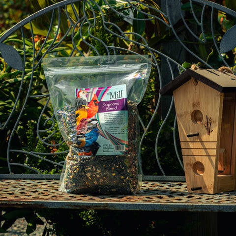 A clear plastic bag of Mill Supreme Blend birdseed sits on a metal garden bench next to a wooden birdhouse, with green garden foliage in the background.