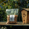 A clear resealable stand-up pouch filled with peanuts sits on a metal garden bench outdoors, next to a wooden birdhouse. The pouch has a white label reading 'Peanut Pits & Skin' with a small bird illustration.