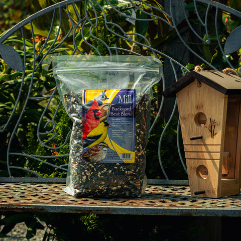 A bag of Mill Mill Backyard Best Blend birdseed sits on a metal garden bench next to a wooden birdhouse, with a decorative metal trellis and green garden foliage in the background.