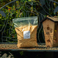 A clear plastic bag filled with coarse yellow-brown granular feed sits on a metal garden bench beside a wooden birdhouse, with green foliage in the background.