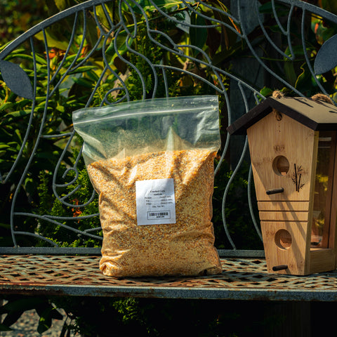 A clear plastic bag filled with coarse yellow-brown granular feed sits on a metal garden bench beside a wooden birdhouse, with green foliage in the background.