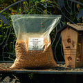 A clear plastic bag filled with corn kernels sits on a metal garden bench outdoors, next to a wooden birdhouse. The bag has a white label reading 'Corn #2 Whole Maized' and the brand 'The Mill'.