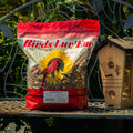 A bag of Birds Luv' Em Peanut Pieces bird feed sits on a black metal garden bench next to a wooden birdhouse. The background shows green garden foliage.