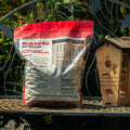 A bag of Birds Luck 'Em Wild Bird Feed sits on a metal garden bench next to a wooden birdhouse, with green foliage in the background.