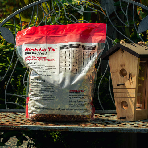 A bag of Birds Luck 'Em Wild Bird Feed sits on a metal garden bench next to a wooden birdhouse, with green foliage in the background.