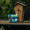 A wooden birdhouse with two circular entry holes stands on a metal garden table next to a bag of Mill Hi-Energy Bird Seed Mix, with a decorative metal fence and greenery in the background.