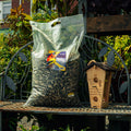 A large clear plastic bag filled with birdseed sits on a metal garden bench next to a wooden birdhouse, with garden foliage and a house in the background.