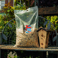A large clear plastic bag filled with birdseed sits on a metal garden bench next to a wooden birdhouse. The bag has a visible The Mill brand label, and the background shows a lush green garden.