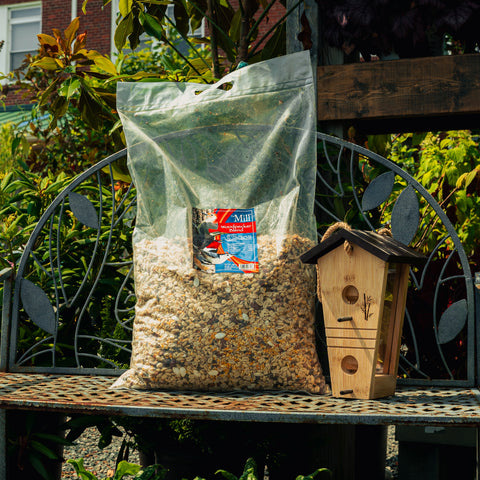 A large clear plastic bag filled with birdseed sits on a metal garden bench next to a wooden birdhouse. The bag has a visible The Mill brand label, and the background shows a lush green garden.