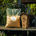 A large clear plastic bag filled with yellow-brown corn kernels sits on a metal garden bench beside a wooden birdhouse, with leafy green plants in the background.