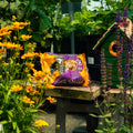 A bag of suet balls on a rustic wooden table in a garden setting, surrounded by yellow flowers, green foliage, and a small green birdhouse.