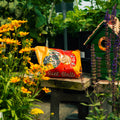 A bag of Hot Pepper Suet Balls placed on a wooden table in a garden setting, surrounded by yellow flowers, green foliage, and a small green birdhouse.