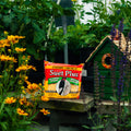 A bag of Cherry Crunch Suet Plus placed on a wooden table in a garden setting with flowers, green plants, and a small green birdhouse.
