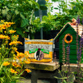 A bag of Suet Plus mealworms and nuts sits on a wooden table in a garden setting, with yellow flowers on the left and a green birdhouse on the right.
