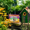 A package of Suet Plus nuts and berry sits on a wooden table in a garden setting, styled with a green birdhouse and plants in the background.