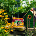 A bag of Suet Plus Peanut Blend  placed on a wooden table in a garden setting, surrounded by yellow flowers, green foliage, and a small green birdhouse.