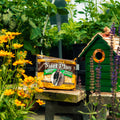A bag of Woodpecker Blend Suet Plus bird food sits on a wooden table in a garden setting, next to a green birdhouse with flowers in the foreground.