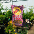 A large purple bag of Birds Luv 'Em Wild Bird Feed stands on a table in a greenhouse, surrounded by green plants and yellow flowers.