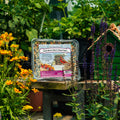 A clear plastic tray of Fresh Berry Nut & Seed Cake bird feed by Pine Tree Farms, displayed on a wooden table in a garden setting with flowers and a birdhouse.