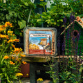A clear plastic clamshell package of Pine Tree Farmer Peanut Butter Sweet Cake sits on a wooden table in a garden setting, surrounded by green foliage, yellow flowers, and a small birdhouse.