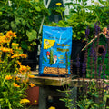 A bag of Woodpecker Suet Nuggets bird feed placed on a wooden table in a garden setting with flowers, plants, and a small birdhouse in the background.