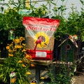 A bag of Birds Luv 'Em Sunflower Meats Seeds displayed on a wooden stand in a garden nursery setting, surrounded by plants, flowers, and a birdhouse.