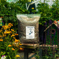 A 5 lb bag of Thistle Seed displayed on a wooden garden table surrounded by green foliage, yellow flowers, and a small green birdhouse.