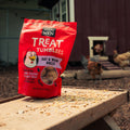 A red bag of Happy Hen Treat Tumblers Oat & Worm Balls chicken treat stands on a wooden table outdoors on a farm, with chickens visible in the background near a barn.