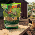 A large bag of Nutrena Chicken Hi Pro Scratch poultry feed is placed on a wooden table in an outdoor backyard setting, with chickens visible in the background near a shed.