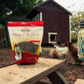 A bag of Durvet Fancy Flock Mealworm & Cricket Blend chicken feed is displayed on a wooden table in a backyard setting with chickens and a chicken coop in the background.