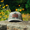 A camouflage trucker cap with a brown leather patch reading 'The Mill' sits on a stone surface outdoors, with yellow flowers and green foliage blurred in the background.