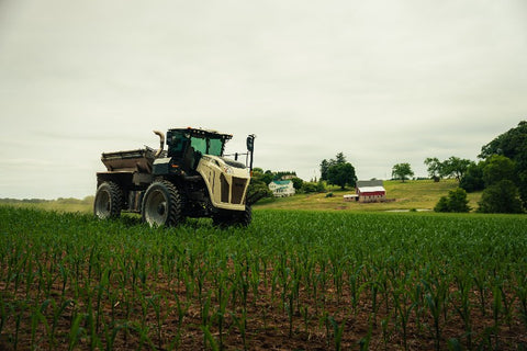 A large agricultural tractor is shown driving across a green field under an overcast sky, with farm buildings and trees in the background.