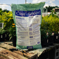 Bag of Coast of Maine Raised Bed Mix on a wooden table with gardening tools and plants in the background.