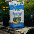 Bag of Espoma Organic Bone Meal plant food on a wooden surface with gardening tools and plants in the background.